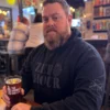 Man wearing Zero Hour blackout tactical hoodie with sleeve graphics, holding a beer at a bar